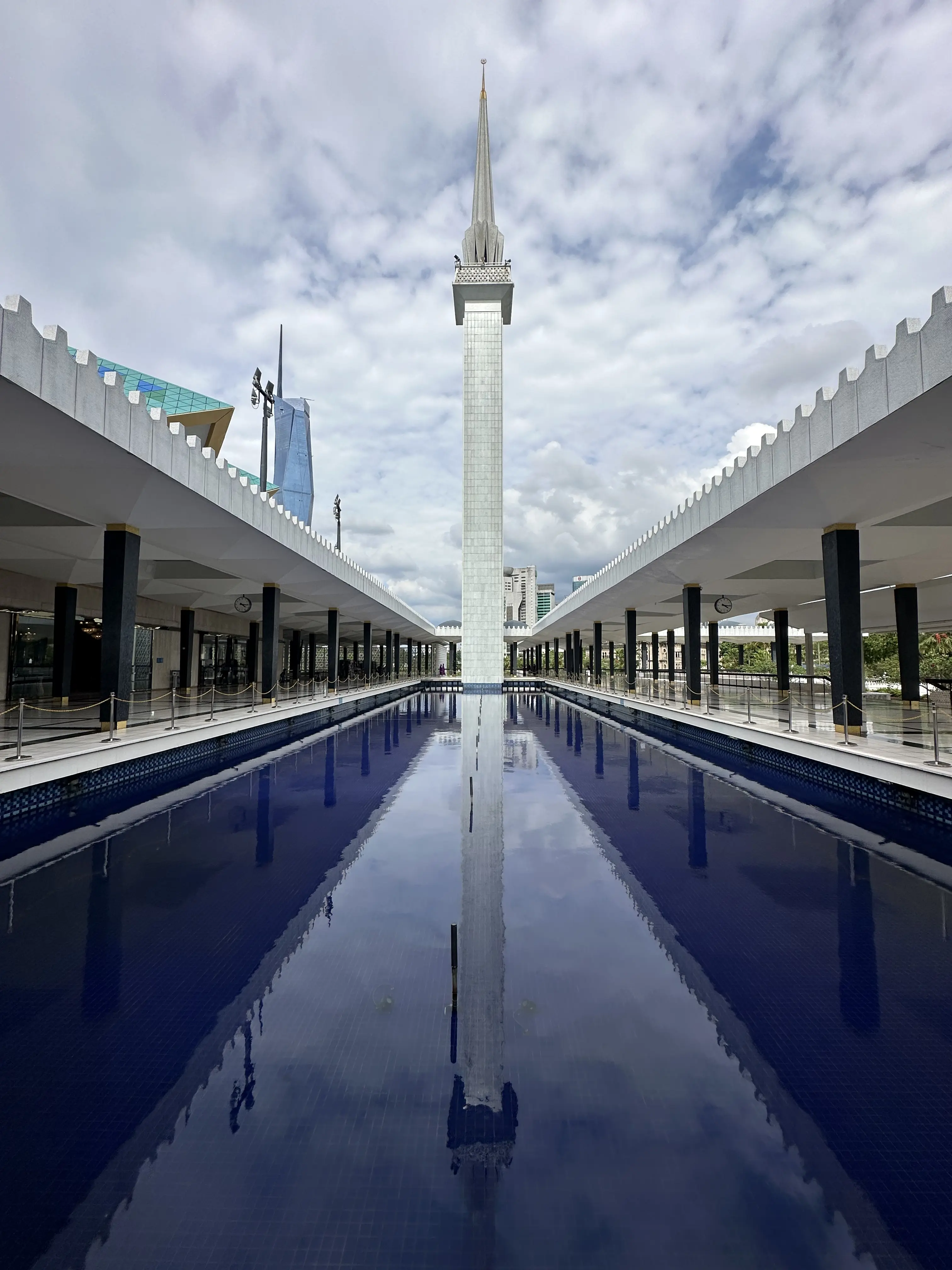 picture in perspective of the minaret of the National Mosque of Malaysia in Kuala Lumpur. On thw way to the minaret there is pool full of bright blue water. On the side of the pool, two corridors with a lot of black columns and a white roof