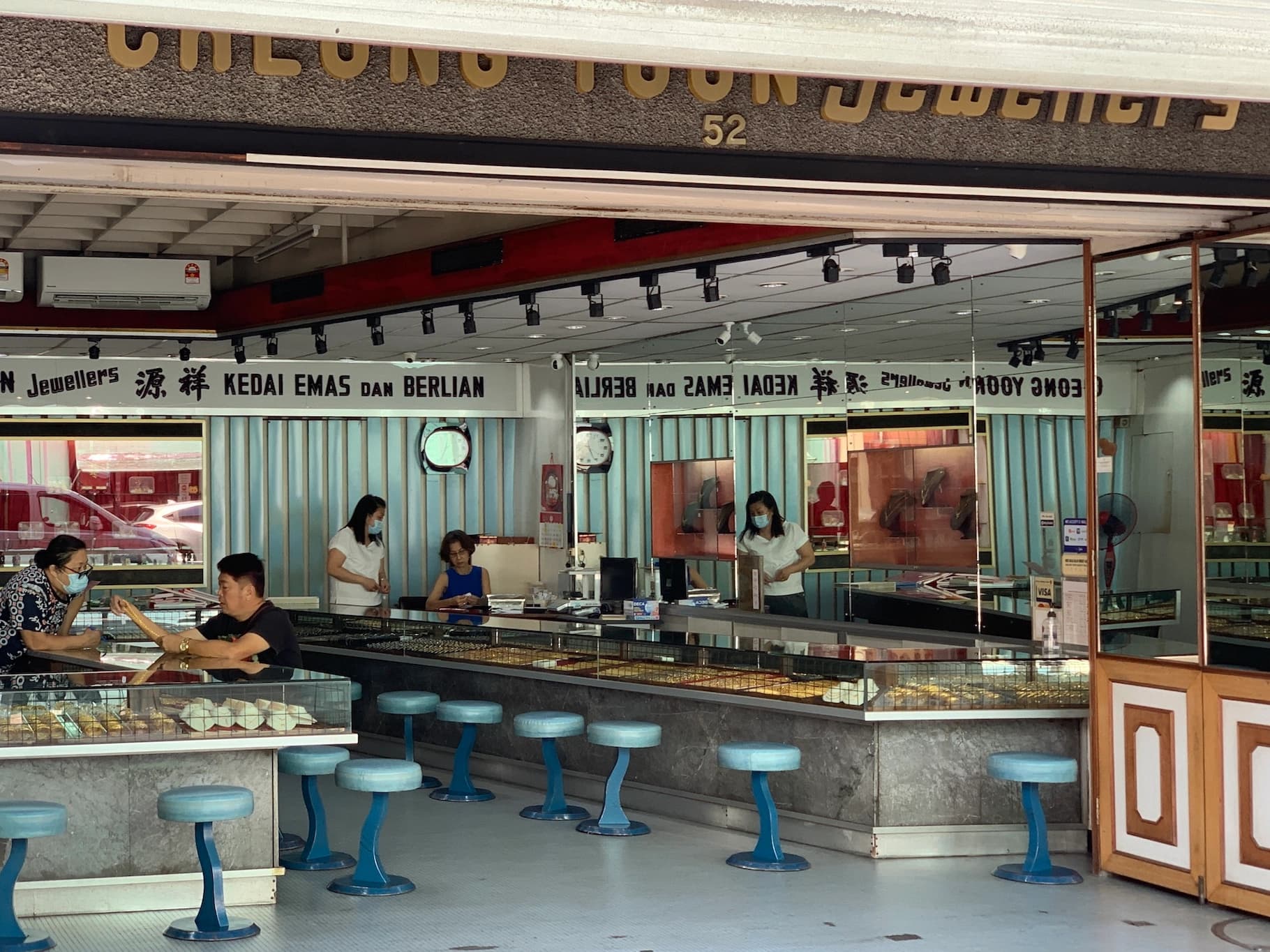 missing image Scene of ordinary life in a gold shop in Malaysia. The shop features two opposite counter, each with a lot of gold items on display. below the counter on the right there a few little, round chairs, coloured in light blue. Inside the shops there a few people. On the right there are two shop assistants talking with each other, while on the left there is a client with a gold necklace on his hand, talking with another shop assitant.