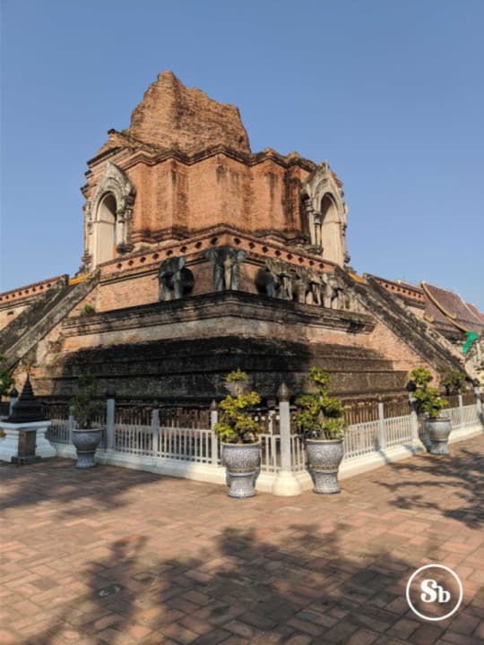 Wat Chedi Luang, Chiang Mai
View of Wat Chedi Luang from one of the corners. The temple is partially in ruins, but you can still make out elephant statues on the sides and the corner. The temple is mainly made up of red bricks. On the upper part, there are two arches, one on each side, of a white-grayish color. In front of the temple, there is a kind of white fence, in front of which there are pots with small plants.