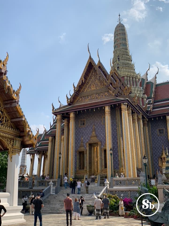 Wat Phra Kaew View of Wat Phra Kaew, home of the Emerald Buddha, in Bangkok. The photo hints at the facade of the palace, with a wall of a bright purple, in front of which stand some beautiful golden columns. The roof is triangular in shape with a red color, and behind the roof a wonder is barely visible.
On the stairs of the palace and in front of it, there are many tourists admiring its beauty, some of whom are also taking pictures.