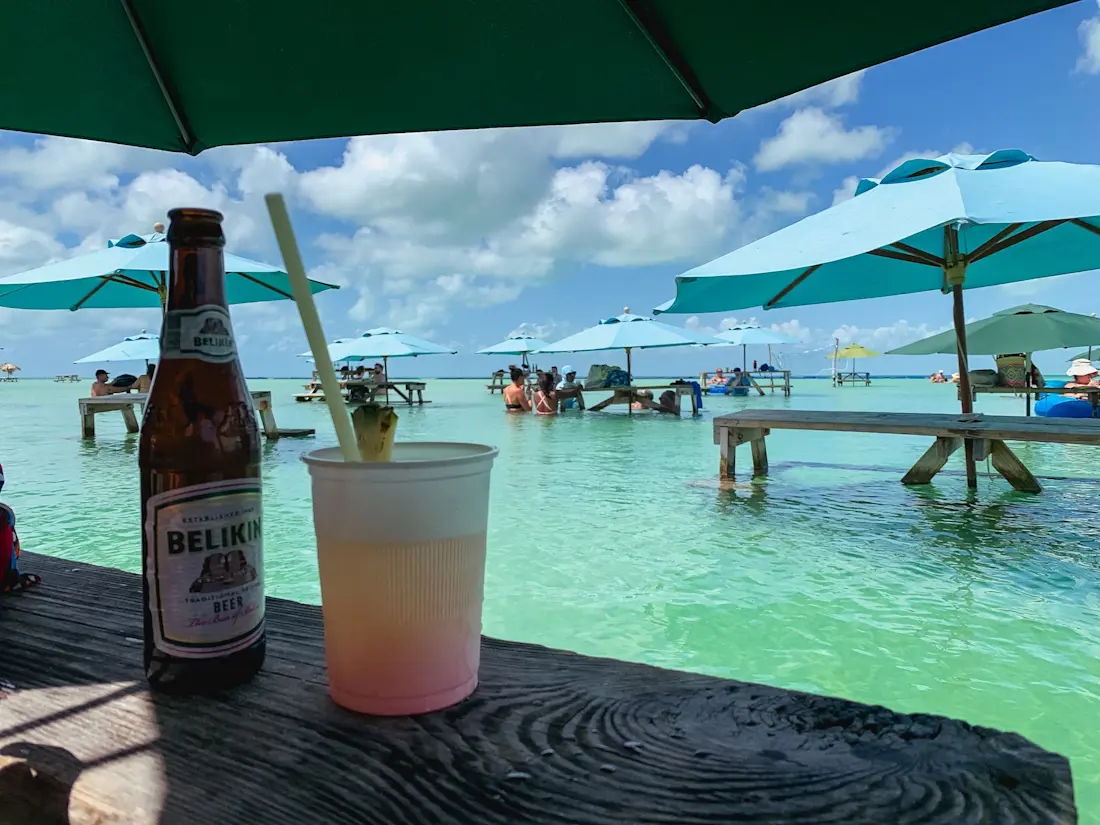 Blue Bayou at Secret Beach Belikin beer bottle and cup with rum punch on a table covered by the shade of an umbrella and Pic-nic tables half submerged in turquoise water and open umbrellas in the background